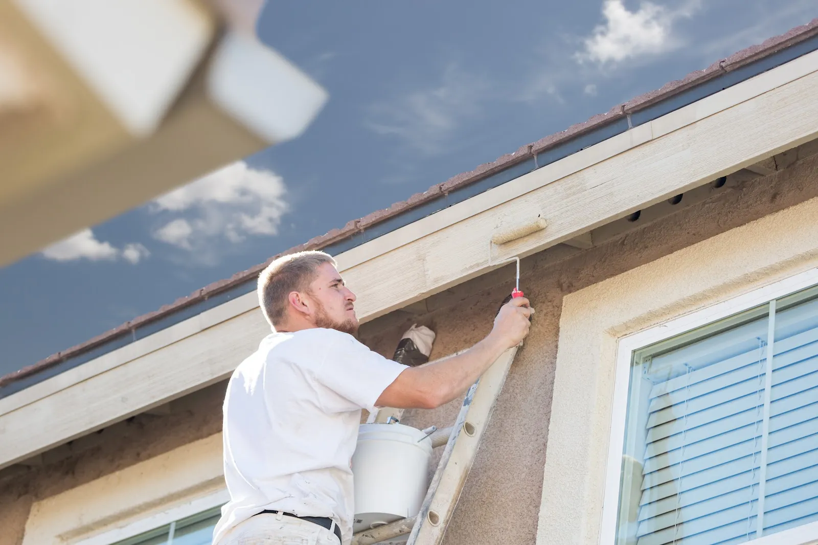 Painter on a ladder rolling white paint onto the fascia of a stucco house