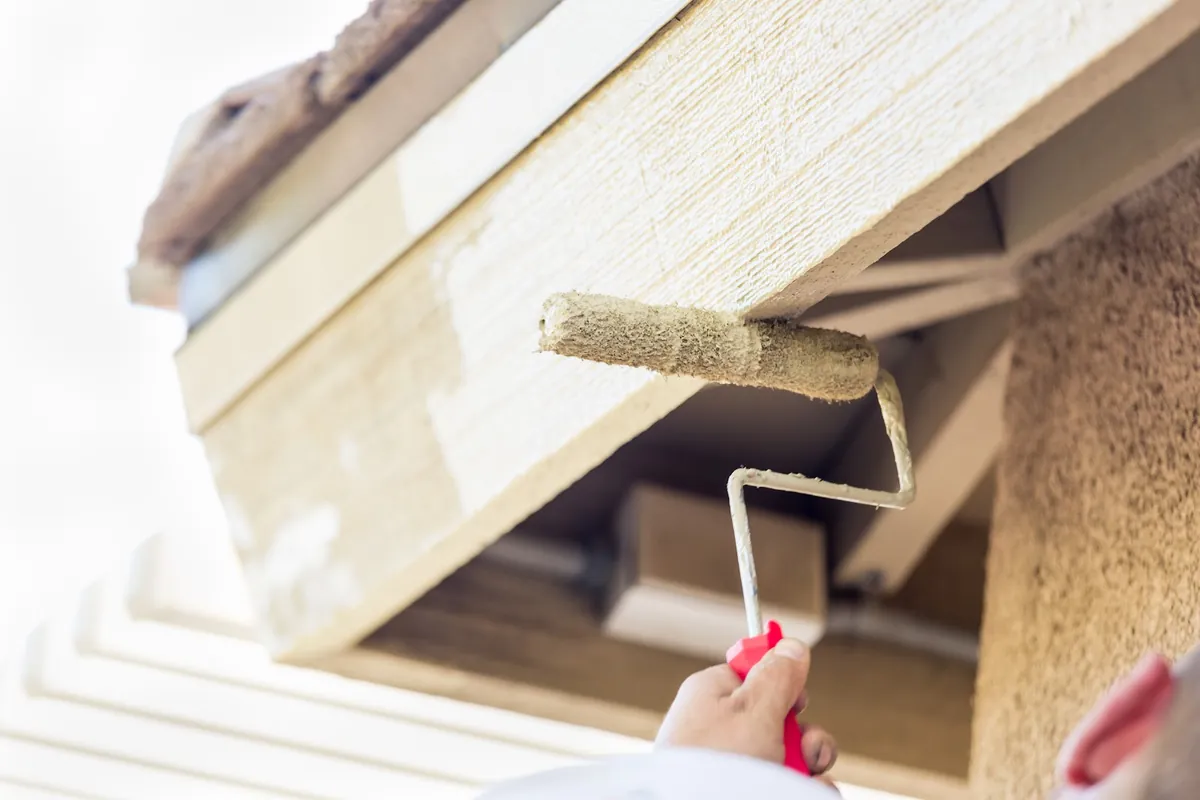 Roller applying fresh paint to wood soffit trim beside a stucco wall